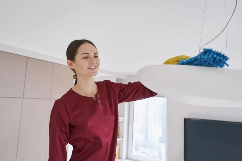Smiling cleaning woman dusting ceiling light fixture