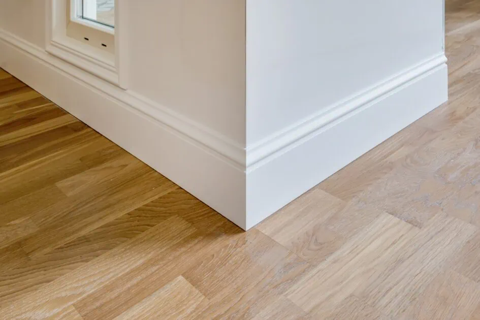 Corner of a clean room with white baseboards and hardwood flooring.