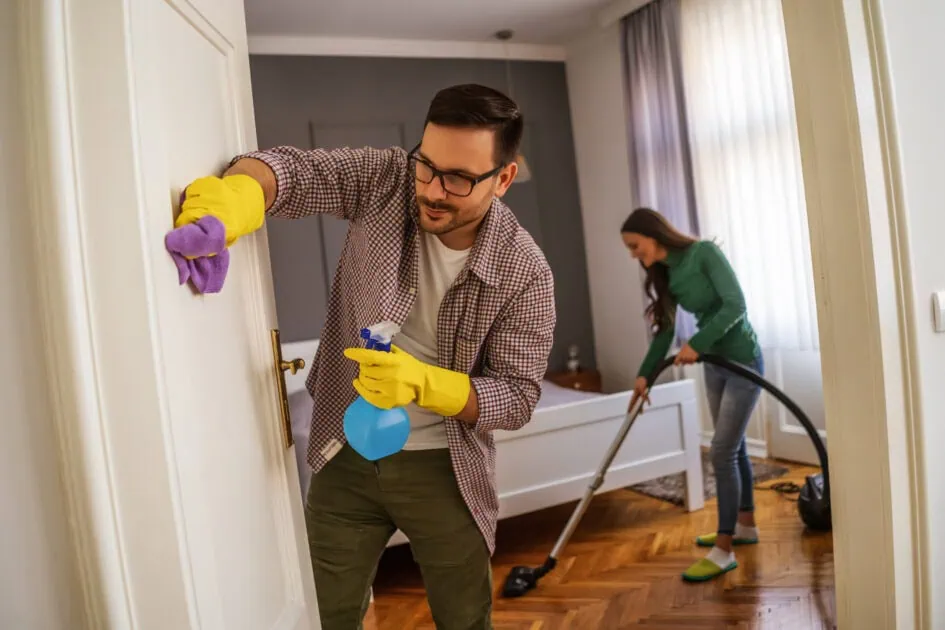 Couple spring cleaning their home—vacuuming floor and wiping door with gloves and spray bottle